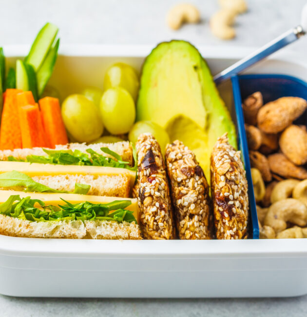 School healthy lunch box with sandwich, cookies, nuts, fruits and avocado on a white background.